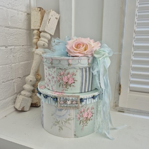 May include: Two stacked round boxes featuring a light blue and white floral pattern with pink rose bouquets. The top box is adorned with a large pale pink fabric rose, light blue tulle, and lace ribbons. A rectangular rhinestone buckle sits on the lower box.