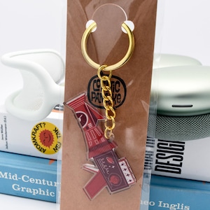 May include: A red and maroon acrylic keychain with a gold chain and ring. The keychain features a rocket ship design with the text "COSMIC PANCAKE". The keychain is attached to a brown card.