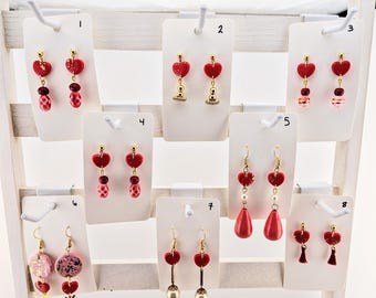 May include: Ten pairs of red heart-shaped earrings with gold accents displayed on a white card stand. The earrings feature various designs, including hearts, beads, and tassels.