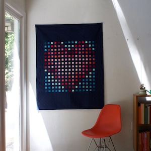 Può includere: Una trapunta blu navy con un motivo a griglia di piccoli quadrati colorati nelle tonalità del rosso, rosa e blu. La trapunta è appesa a una parete bianca. Una sedia arancione è sotto.
