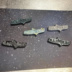 Peut inclure: Plusieurs épingles métalliques avec le mot "ILLENIUM" dans une police stylisée. Les épingles sont de différentes couleurs, dont l'argent, le noir et le sarcelle, et sont disposées sur un fond sombre parsemé de points blancs.