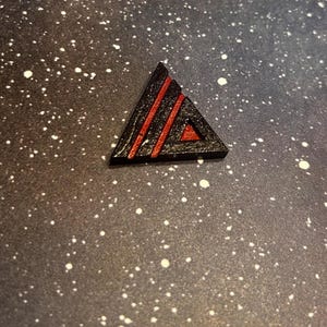 May include: A black triangular wooden shape with red stripes. The shape is on a dark gray background with white speckles.
