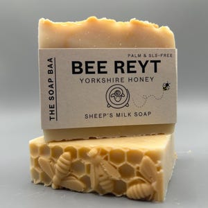 May include: A rectangular bar of light beige soap with a textured honeycomb and bee pattern on the bottom. A paper label wraps around the centre with the text: THE SOAP BAA, BEE REYT, YORKSHIRE HONEY, SHEEP'S MILK SOAP, and PALM & SLS-FREE.