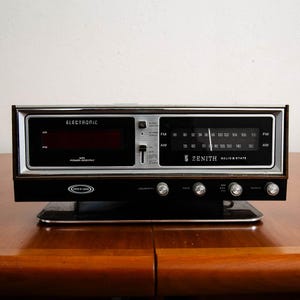 May include: A vintage Zenith Solid State electronic clock radio featuring a dark wood-grain finish, a black front panel with a digital display, and an analog AM/FM tuner dial. The unit includes silver control knobs for volume, tone, and tuning.