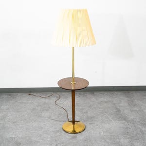 May include: A floor lamp featuring a cream-colored pleated lampshade, a slender brass central pole, and a circular dark wood side table integrated into the stand. The lamp rests on a circular brass base and includes a thin brown electrical cord.
