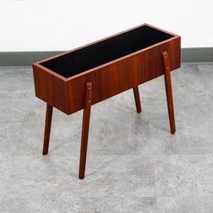 May include: A mid-century modern rectangular planter made of wood, featuring a rich brown finish. The planter has a black interior and is supported by four angled wooden legs. The legs are attached with small metal accents.