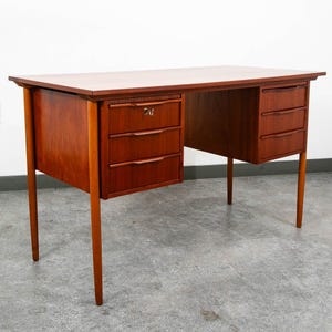 May include: A wooden desk with a rectangular top and four tapered legs. The desk features two sets of drawers, with three drawers on each side. The top left drawer includes a visible metal keyhole. The wood has a warm, medium-brown finish.