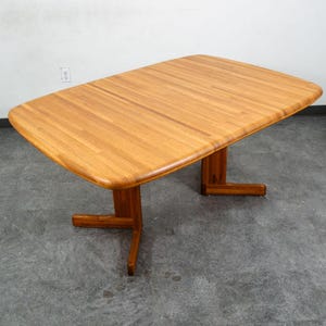 May include: A rectangular wooden dining table with a light brown finish and visible wood grain. The tabletop features rounded corners and is supported by two T-shaped pedestal bases, each with a horizontal foot extending outward on both sides.