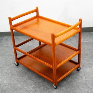 Peut inclure: Chariot de service en bois de style moderne du milieu du siècle. Il comporte deux étagères et des poignées. Il repose sur des roulettes. Le bois est d'un ton brun-rougeâtre chaud. Mesures: 76 cm de long, 46 cm de large, 76 cm de haut.