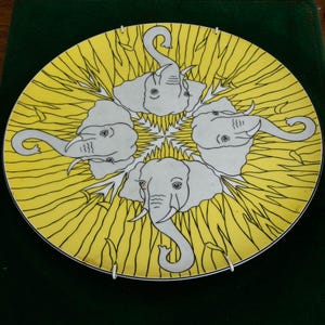 May include: A round decorative plate featuring a bright yellow background with a repeating pattern of four white elephant heads arranged in a circular formation. Each elephant is outlined in black, with trunks pointing outward and tusks visible.