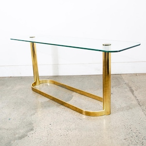 以下が含まれることがあります： 長方形のガラス天板と、ゴールドカラーの金属フレームが特徴的なモダンなコンソールテーブル。 フレームは曲線的なベースと円筒形の脚を備え、洗練されたミニマリストデザインを演出。 テーブルの長さは約122cmです。