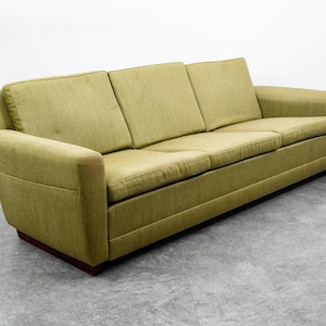 May include: A three-cushion sofa with a light green fabric. The sofa has a low profile with a dark brown wooden base and rectangular arms. The cushions and arms are a solid color, and the sofa is set against a white wall.