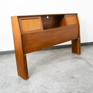 May include: A wooden headboard featuring a smooth, medium-brown finish with two rectangular cane-paneled sections on the upper left and right sides. A central open storage shelf is positioned between the cane panels with a circular hole.