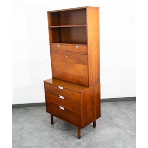 May include: A tall wooden secretary desk featuring a warm brown finish, an upper open shelf, two small drawers, a drop-down writing surface, and three lower storage drawers. The piece stands on four tapered legs and includes silver-toned hardware.