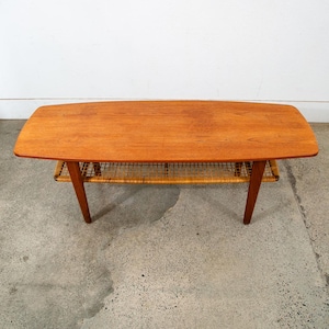 May include: A mid-century modern coffee table with a rectangular, light brown wooden top and a woven rattan shelf. The table has angled wooden legs and is set against a neutral background. The table measures approximately 48 inches long.