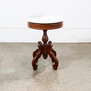 May include: A round side table featuring a white marble top with grey veining. The table has a dark brown wooden base with a central pedestal column supported by four curved, ornate legs that extend outward from the base of the central post.