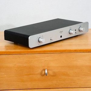 May include: A black and silver audio amplifier with multiple knobs and a headphone jack. The amplifier has the word "Classic" printed on the front. The device is rectangular and sits on a wooden surface.