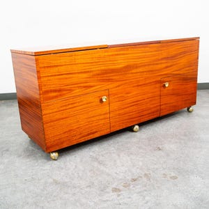 May include: A rectangular wooden sideboard featuring a rich mahogany-toned grain, two cabinet doors, and a top surface. The piece is supported by four small, spherical brass-colored casters and includes two small, round brass-colored knobs.