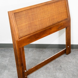 May include: A rectangular wooden headboard featuring a central panel of woven cane material. The frame is constructed from medium-toned wood with a smooth finish, and the structure includes two vertical legs with visible metal hardware bolts.