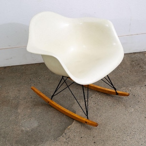 May include: A mid-century modern rocking chair with a white, speckled plastic seat. The chair has a black wire base and wooden rockers. The design is minimalist and classic, suitable for a living room or nursery.