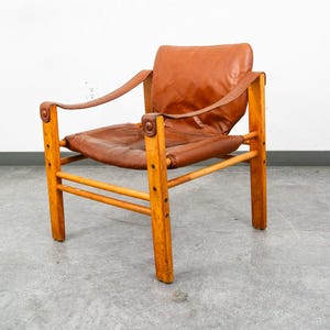 May include: A lounge chair featuring a light brown wooden frame with vertical supports and horizontal crossbars. The seat and backrest are composed of a single piece of tan leather that is suspended between the armrests and secured with circular studs.