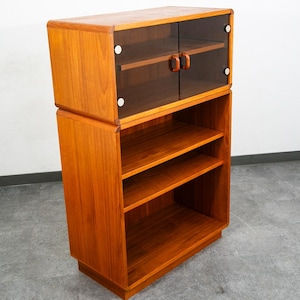 May include: A tall wooden cabinet featuring a warm brown wood grain finish. The upper section includes two glass doors with small wooden handles and metal hinges. The lower section consists of an open storage area with two fixed wooden shelves.