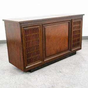 May include: A rectangular wooden media console featuring a smooth top surface and a central solid panel flanked by two vertical speaker sections. Each speaker section has a decorative lattice grille pattern over a dark fabric backing.