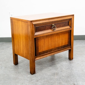 May include: A wooden nightstand featuring a rectangular top, one drawer with a brass ring pull, and a lower cabinet door. The piece is constructed from medium-toned wood with darker wood grain accents on the drawer front and cabinet panel.