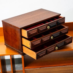 May include: A wooden jewelry chest with three pull-out drawers. Each drawer front features a geometric carved pattern with dark brown accents and a small gold-toned knob. The interior of each drawer is lined with a vibrant red velvet fabric.