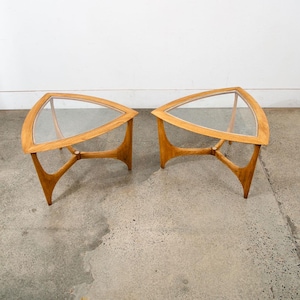 May include: Two mid-century modern side tables. Each table has a triangular glass top with a wooden frame. The wooden legs are curved and connected by a horizontal support. The tables are made of light-colored wood.