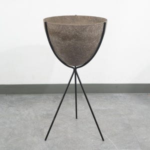 May include: A mid-century modern planter with a textured, brown bowl supported by a black, tripod metal stand. The bowl has a wide, open top and a rounded shape. The stand's legs are thin and angled.