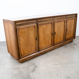 May include: A mid-century modern wooden cabinet with four doors and a light-coloured top. The cabinet is made of wood with a visible grain pattern and dark brown trim. The doors have small, dark handles.