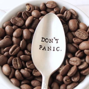 Peut inclure: Une cuillère en argent gravée des mots "Ne paniquez pas", entourée de grains de café dans une tasse en céramique blanche.