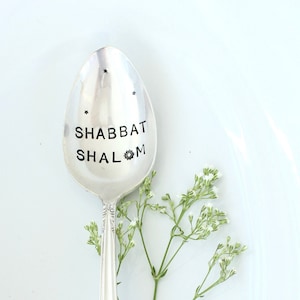 May include: A silver spoon with the words "SHABBAT SHALOM" stamped on the bowl. The spoon is decorated with small star-shaped designs and a floral sprig. The spoon is resting on a white surface.