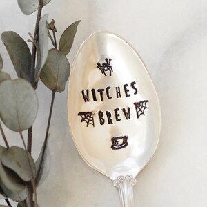 Puede incluir: Una cuchara de plata con las palabras "Witches Brew" grabadas. La cuchara tiene un diseño de araña y una taza de té.