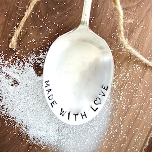 May include: A silver spoon engraved with the words "Made with Love" and a small heart symbol. The spoon is lying on a wooden surface with white sugar crystals scattered around it.