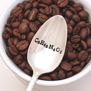 Puede incluir: Una cuchara de plata con la fórmula química de la cafeína, C8H10N4O2, grabada en ella. La cuchara está apoyada en una taza blanca llena de granos de café.