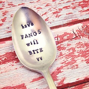 以下が含まれることがあります： 「have FANGS will BITE」と刻まれた銀のスプーン。スプーンは赤と白の木製表面の上に置かれています。