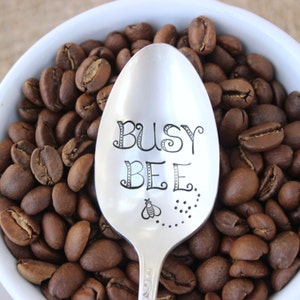 May include: A silver spoon with the words "Busy Bee" engraved on it, surrounded by coffee beans.
