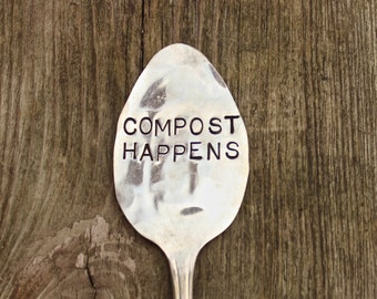 May include: A silver spoon with the words "COMPOST HAPPENS" engraved on it. The spoon is sitting on a wooden surface.