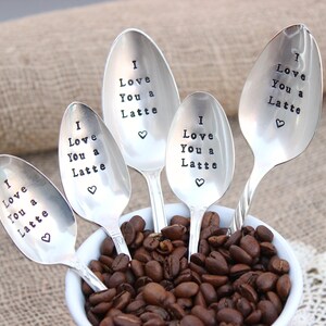 May include: Five silver spoons with the phrase "I Love You a Latte" engraved on them. The spoons are arranged in a white bowl filled with coffee beans.