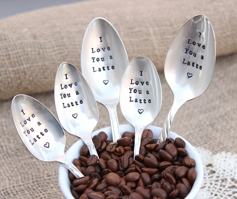 May include: Five silver spoons with the phrase "I Love You a Latte" engraved on them. The spoons are arranged in a white bowl filled with coffee beans.