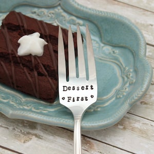 May include: Silver fork with the words "Dessert First" engraved on the handle. The fork is resting on a blue and white ceramic plate with a piece of chocolate cake on the plate.