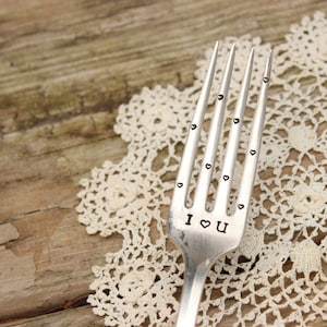 May include: A silver fork with the words "I ♡ U" stamped on the tines. Small heart-shaped designs are stamped along the tines. The fork is set on a cream-colored lace doily and a wooden surface.