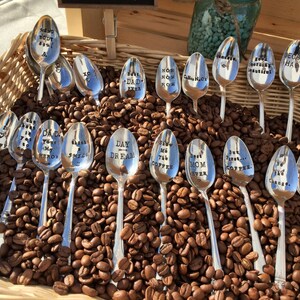 May include: A collection of silver spoons with various coffee-themed sayings engraved on them, displayed in a wicker basket filled with coffee beans.