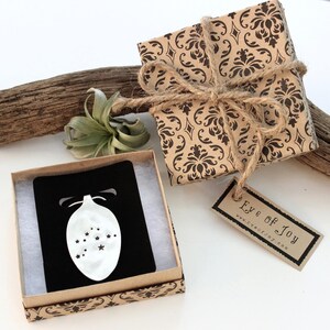 May include: A gift box wrapped in brown paper with a black damask pattern, tied with twine. A small box with a black card and a white ornament design is open next to it. The tag reads "Eye of Joy".