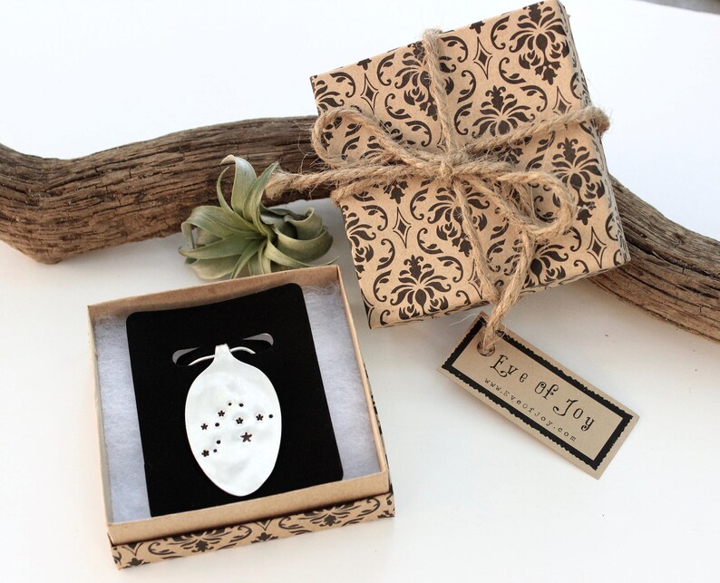 May include: A gift box wrapped in brown paper with a black damask pattern, tied with twine. A small box with a black card and a white ornament design is open next to it. The tag reads "Eye of Joy".
