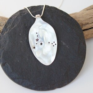 May include: A silver spoon pendant necklace with a constellation design. The spoon-shaped pendant features a polished, reflective surface with small star-shaped and dot-shaped cutouts. The necklace is displayed on a dark gray, round stone.