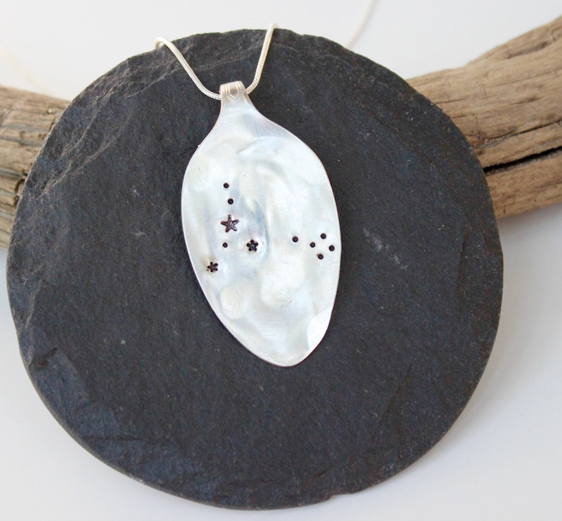 May include: A silver spoon pendant necklace with a constellation design. The spoon-shaped pendant features a polished, reflective surface with small star-shaped and dot-shaped cutouts. The necklace is displayed on a dark gray, round stone.