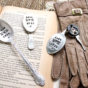 May include: Three silver spoons with the words "Best Mom Ever" stamped on them. The spoons are on a book and a brown leather glove with a key chain.